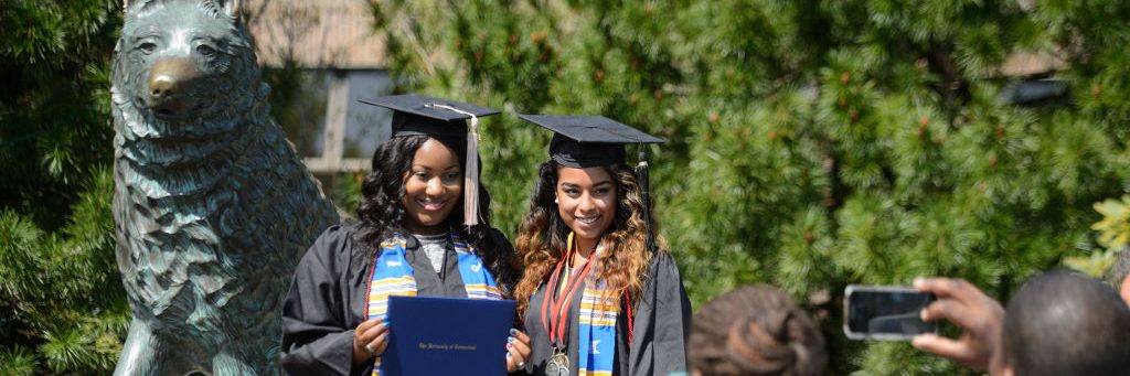 two females of african descent in commencement regalia celebrating their commencement in front of the husky statue.